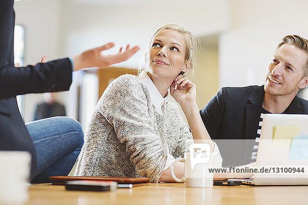 Colleagues looking at businesswoman talking while sitting on table in office