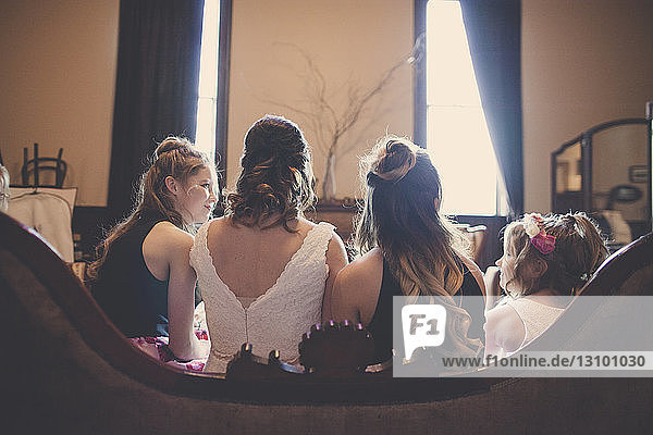 Rückansicht der Braut mit Brautjungfern und Blumenmädchen auf dem heimischen Sofa sitzend