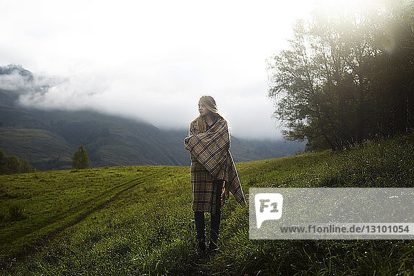 Nachdenkliche  in eine Decke gehüllte Frau schaut weg  während sie im Winter auf einem Grasfeld steht