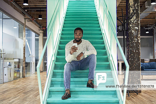 Geschäftsmann benutzt Mobiltelefon  während er im Büro auf Stufen sitzt