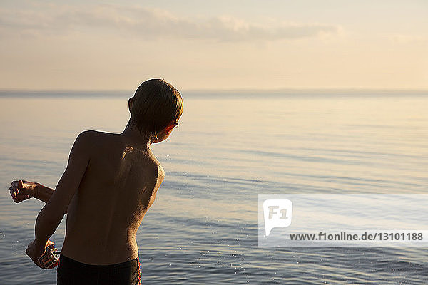 Rear view of wet boy skipping stones by sea during sunset