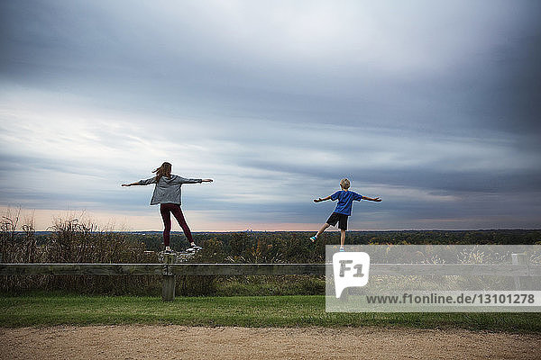 Teenage girl and boy standing on one leg on wooden fence against sky