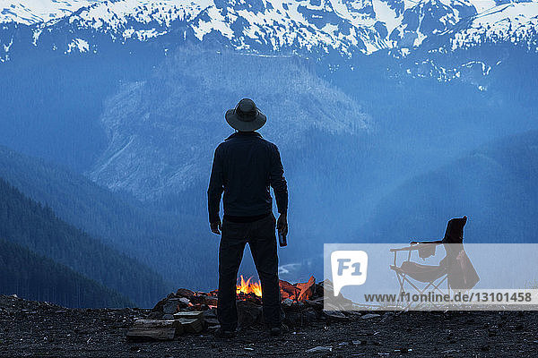 Mann steht am Lagerfeuer vor schneebedeckten Bergen
