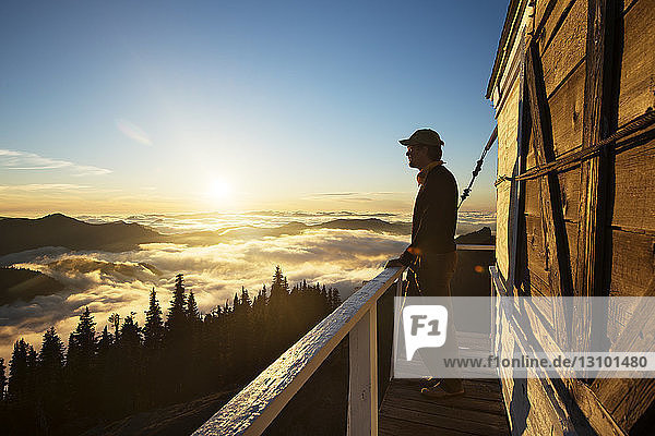 Side view of man looking at mountain view from log cabin during sunset