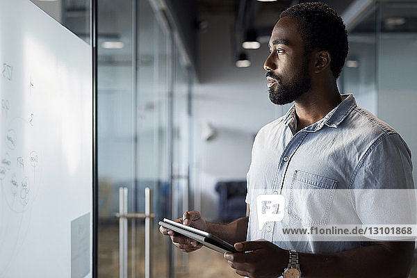 Nachdenklicher Geschäftsmann benutzt digitales Tablet  während er im Bürokorridor steht