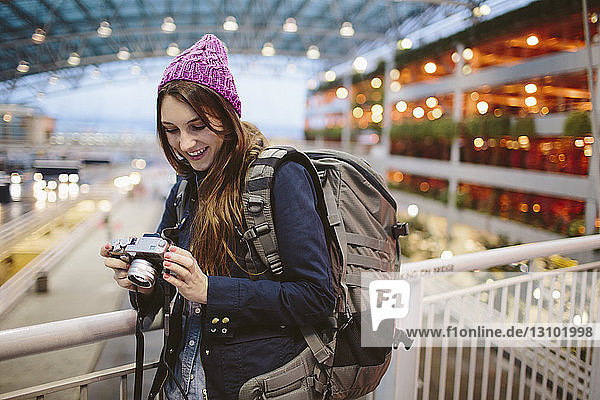 Lächelnde Frau mit Rucksack schaut in die Kamera  während sie sich am Geländer im Flughafen anlehnt