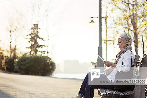 Seitenansicht einer älteren Frau  die einen Tablet-Computer benutzt  während sie auf einer Bank am Fußweg sitzt