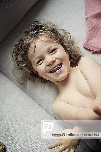 Hochwinkelporträt eines glücklichen Jungen ohne Hemd  der zu Hause auf dem Sofa liegt