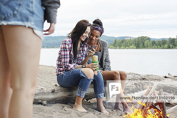 Freundinnen benutzen ein Smartphone  während sie am Lagerfeuer am Fluss auf einem Baumstamm sitzen