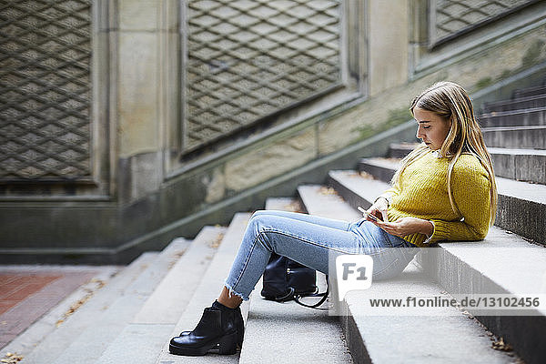 Seitenansicht einer Frau in voller Länge  die ein Smartphone benutzt  während sie auf einer Treppe im Park sitzt