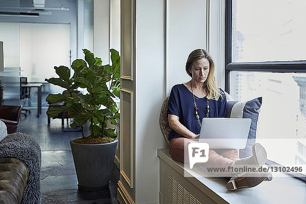 Businesswoman using laptop computer while sitting on window sill in office