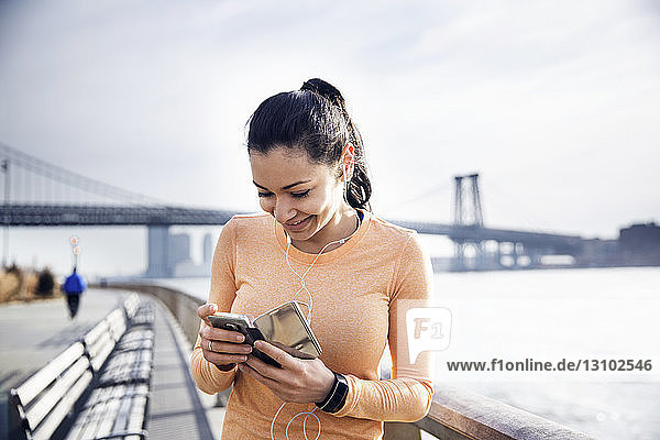 Glückliche Sportlerin hört Musik über Smartphone mit Williamsburg Bridge im Hintergrund