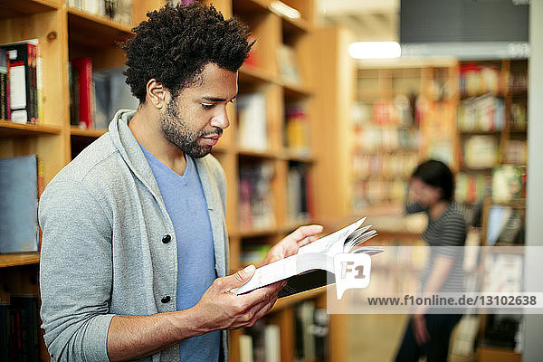 Man reading book in library with woman standing in background