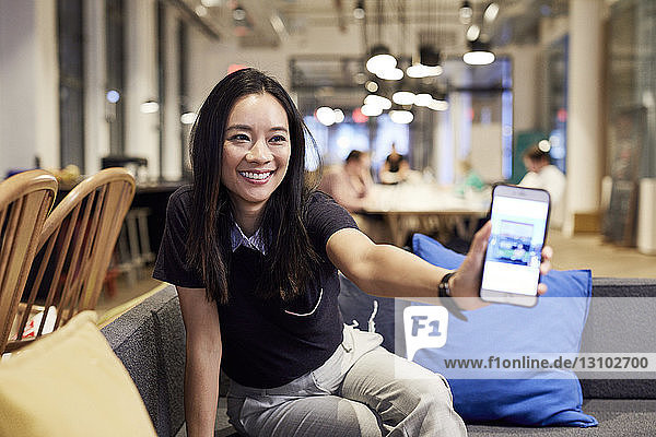 Lächelnde Geschäftsfrau zeigt Mobiltelefon  während sie im Büro auf dem Sofa sitzt