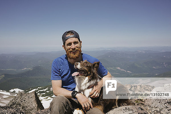 Portrait of man with dog sitting on mountain against sky