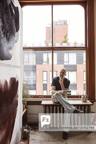 Thoughtful artist holding coffee cup while sitting by window at workshop