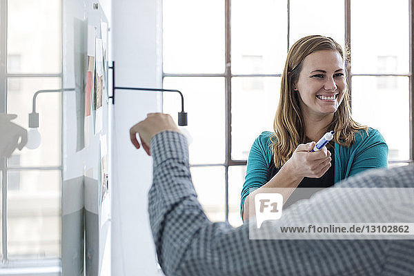 Lächelnde Geschäftsfrau plant mit Kollegin  während sie im Büro am Whiteboard steht