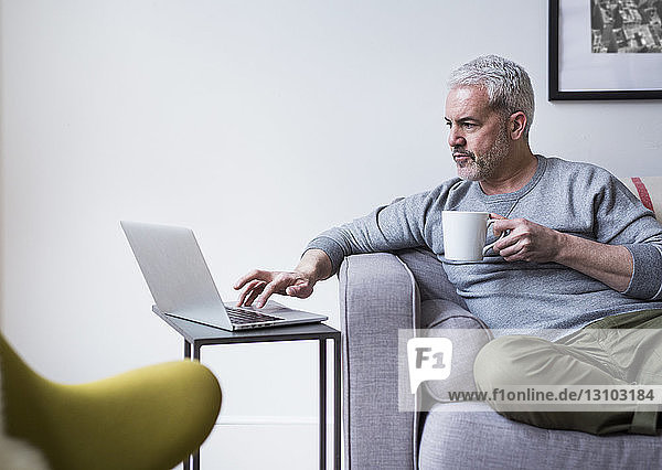 Mature man using laptop while holding coffee mug at home