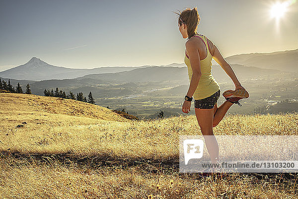 Side view of woman exercising on grassy mountain against sky during sunny day