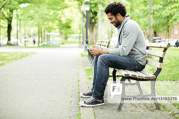 Seitenansicht eines glücklichen Mannes  der ein Smartphone benutzt  während er auf einer Parkbank am Fußweg sitzt