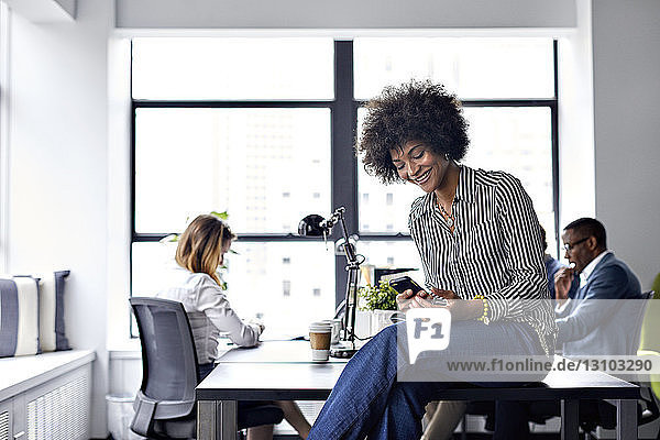 Glückliche Geschäftsfrau benutzt Smartphone  während Kollegen im Hintergrund arbeiten
