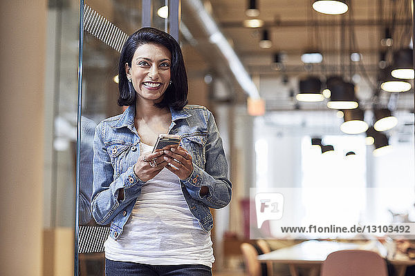 Glückliche Geschäftsfrau schaut weg  während sie ein Smartphone im Büro hält