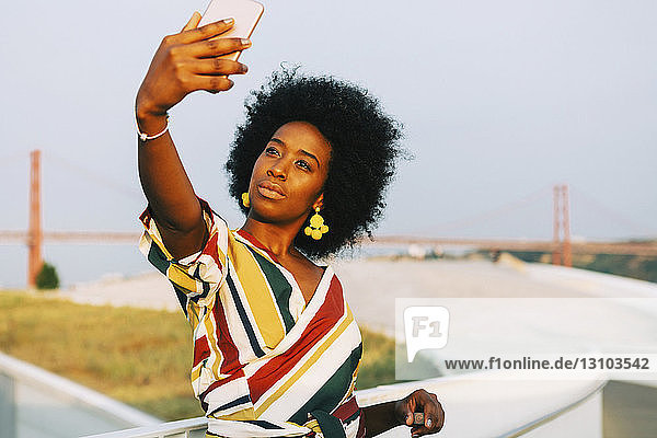 Selbstbewusste junge Frau macht ein Selfie mit der Brücke 25 de Abril im Hintergrund  Lissabon  Portugal