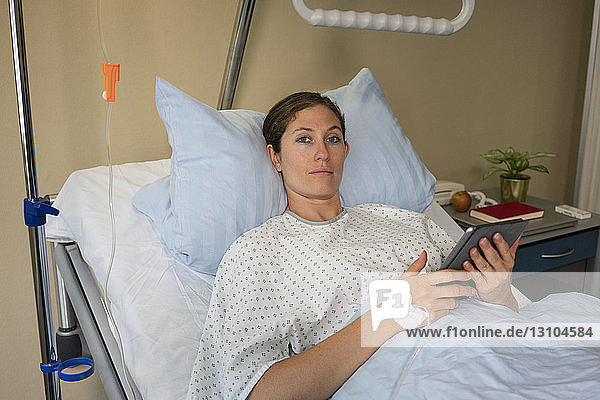 Portrait serious female patient with digital tablet  resting and recovering in hospital room