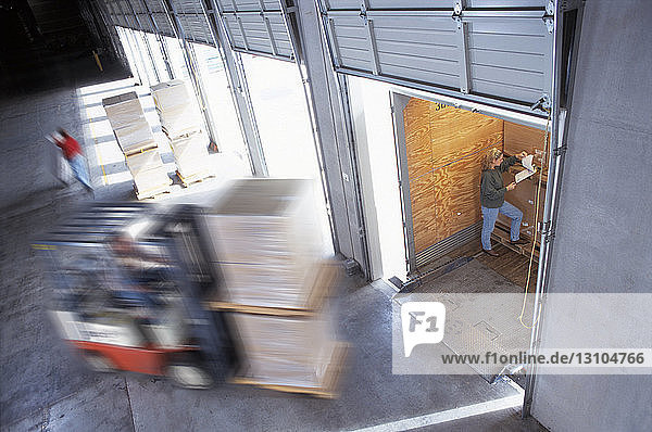 Employees loading cardboard boxes of products into truck trailers in a distribution warehouse loading dock area  one checking a manifest.