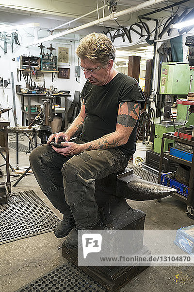 Male Blacksmith checking his phone for messages while sitting on an anvil in his studio.