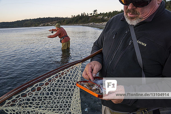 Ein Fliegenfischer-Guide überprüft sein Fliegensortiment auf Lachs und Searun-Küstenschneeforelle  während sein Klient im Hintergrund mit der Fliege fischt.