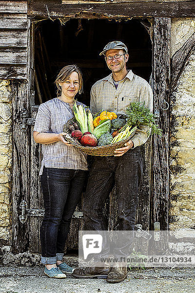Smiling farmer and woman standing in barn door  holding basket with freshly harvested vegetables  looking at camera.