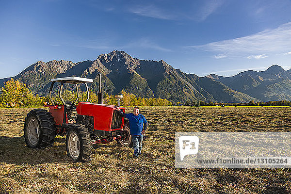 A farmer wearing a blue shirt stands next to an old red tractor in an open field  Pioneer Peak in the background  South-central Alaska; Palmer  Alaska  United States of America