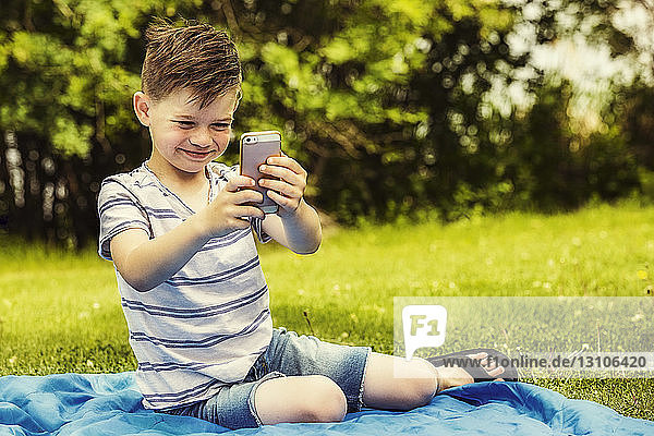 A young boy making silly facial expressions while taking self-portraits with a smart phone in a city park on a warm summer day; Edmonton  Alberta  Canada