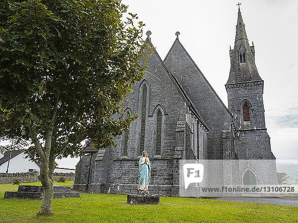 Steinkirche mit Glockenturm und Statue einer betenden Frau im Vordergrund; County Clare  Irland