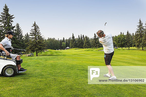 Ein männlicher Golfer schlägt einen Golfball mit einem Wedge auf einem Golfplatz auf das Grün  während ein behinderter Golfer in einem Spezialrollstuhl zusieht; Edmonton  Alberta  Kanada