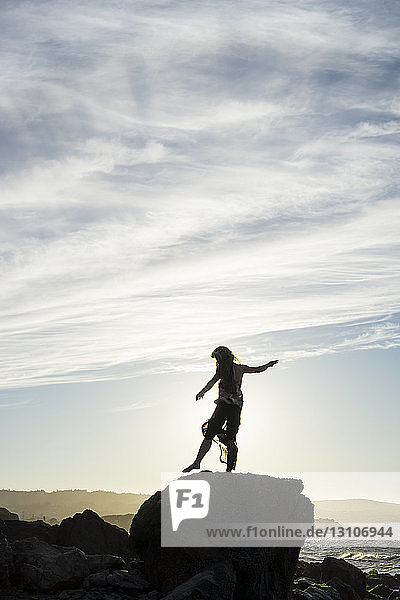 Eine Frau steht auf einem Fuß balancierend auf einem Felsen mit Blick auf die Küste bei Sonnenuntergang  silhouettiert und beleuchtet vom Sonnenlicht; San Mateo  Kalifornien  Vereinigte Staaten von Amerika
