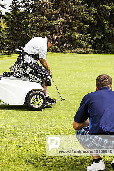 Ein nichtbehinderter Golfer hilft einem behinderten Golfer,  der einen speziellen elektrischen Golfrollstuhl benutzt,  seinen Putt auf dem Grün eines Golfplatzes zu platzieren; Edmonton,  Alberta,  Kanada