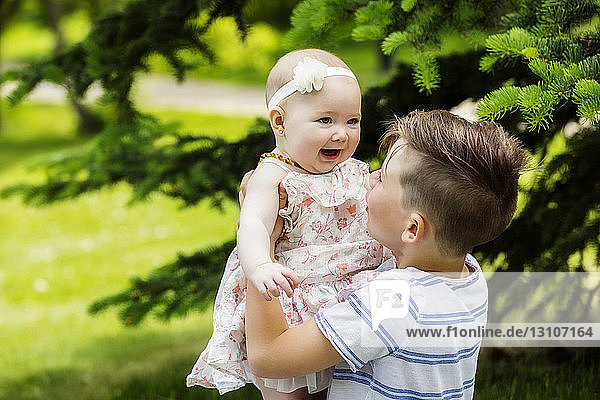 Ein kleiner Junge spielt mit seiner kleinen Schwester und hält sie in einem Stadtpark an einem warmen Sommertag in die Luft; Edmonton  Alberta  Kanada