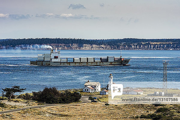 Frachtschiff  das von der Straße von Juan de Fuca über Admiralty Inlet in den Puget Sound einfährt. Der Point Wilson-Leuchtturm befindet sich im Fort Worden Historical State Park in der Nähe der Stadt Port Townsend. Im Hintergrund ist Whidbey Island zu sehen; Washington  Vereinigte Staaten von Amerika