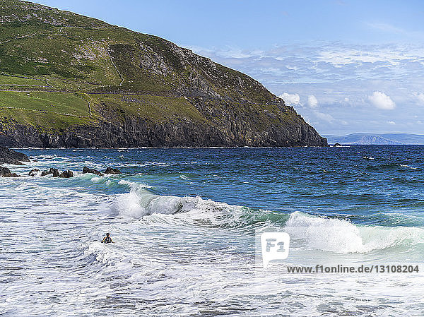 Rugged coastline with cliffs and mountains and a swimmer in the ocean waves in the foreground; Ballyferriter  County Kerry  Ireland