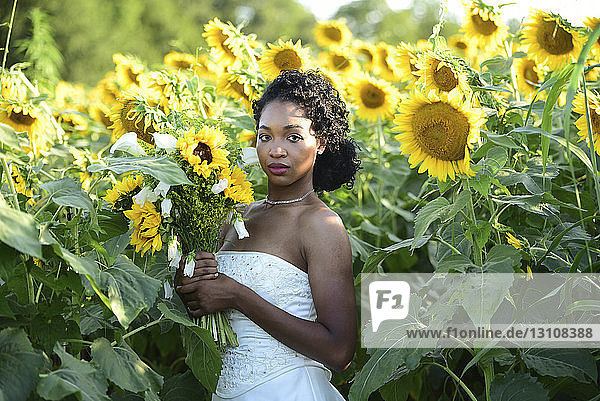 Porträt einer Braut  die einen Blumenstrauss hält  während sie bei Sonnenblumen auf dem Bauernhof steht