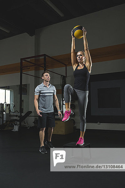 Trainer sieht Frau an  die im Fitness-Studio einen Fitnessball auf einem Hocker stehend hält