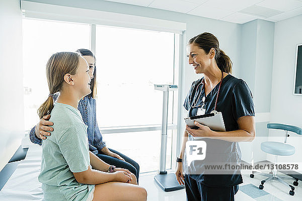 Fröhliches Mädchen schaut den Kinderarzt an  während sie im Krankenhaus neben der Mutter auf dem Untersuchungstisch sitzt