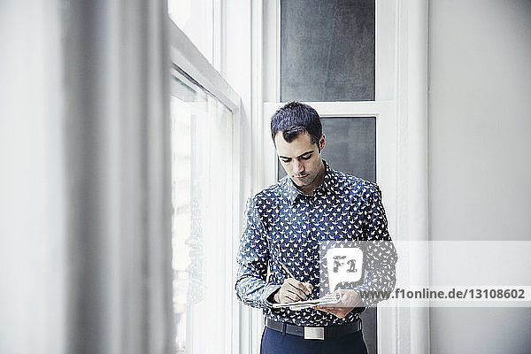 Geschäftsmann schreibt in Buch  während er im Kreativbüro am Fenster steht