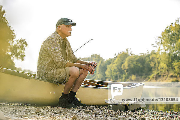 Nachdenklicher älterer Mann sitzt auf einem Boot am Seeufer vor klarem Himmel