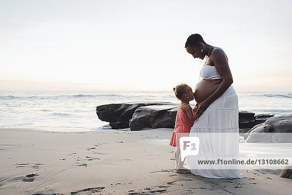 Tochter küsst schwangeren Mutterbauch  während sie bei Sonnenuntergang am Strand steht