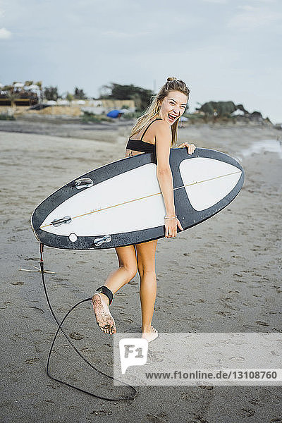 Fröhliche Frau trägt Surfbrett beim Strandspaziergang