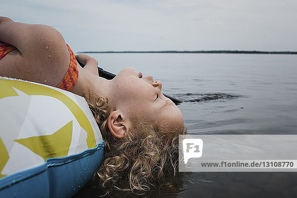 Side view of cute girl with eyes closed lying in inflatable raft on lake against sky