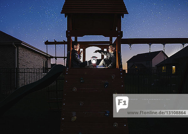 Brüder mit beleuchteter Laterne sitzen auf Außenspielgeräten gegen das Sternenfeld im Park